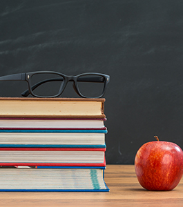 Eyeglasses on top of stacked books on a table next to an apple and in front of a chalkboard
